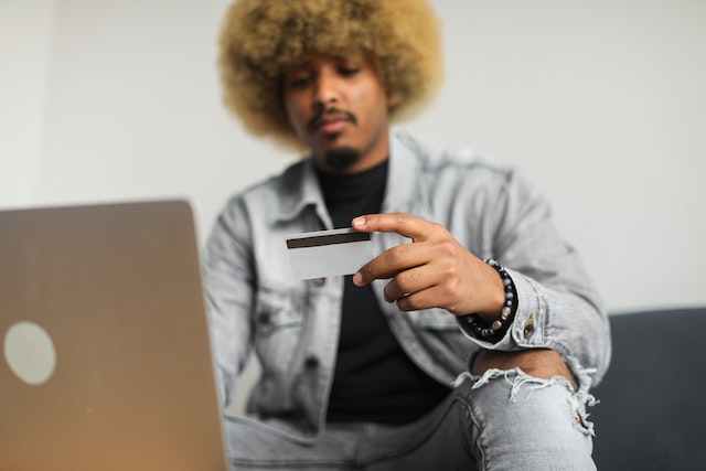 person in denim jacket looking at their credit card while making an online payment on their laptop